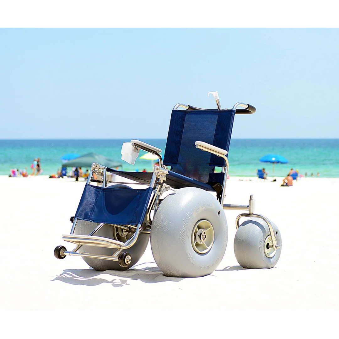 The DeBug All-Terrain Beach Wheelchair with large gray inflatable wheels sits on white sand near the ocean, ideal for outdoor mobility and comfort at the shore, with people and blue umbrellas in the background.